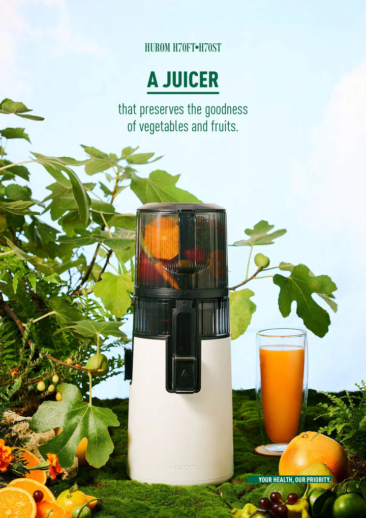 Black and white Hurom H410 slow juicer with fruits and a glass of juice surrounded by greenery