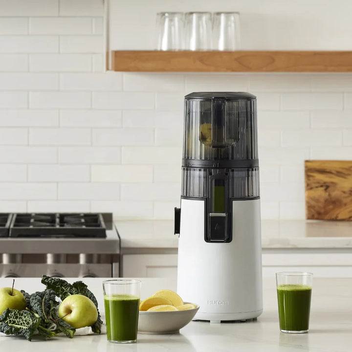 White and black Hurom H410 juicer on a kitchen counter with green juice and fruits.