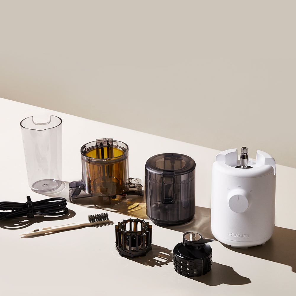 A charcoal black disassembled Hurom H-310A Slow Juicer on a table, with a power cable and brush.