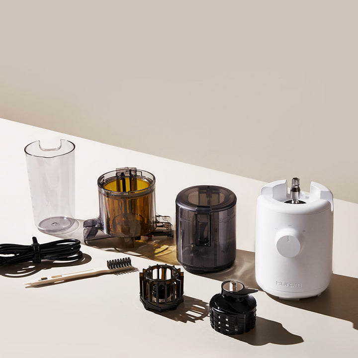 A charcoal black disassembled Hurom H-310A Slow Juicer on a table, with a power cable and brush.