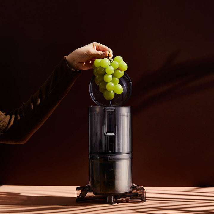 A charcoal black Hurom H-310A Slow Juicer on a table, with grapes being placed into the juicer.