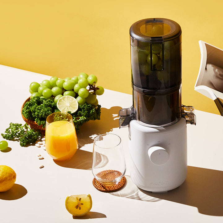 A lavender Hurom H-310A Slow Juicer on a table, with grapes, broccoli, lemons and a glass of lemon juice beside it.