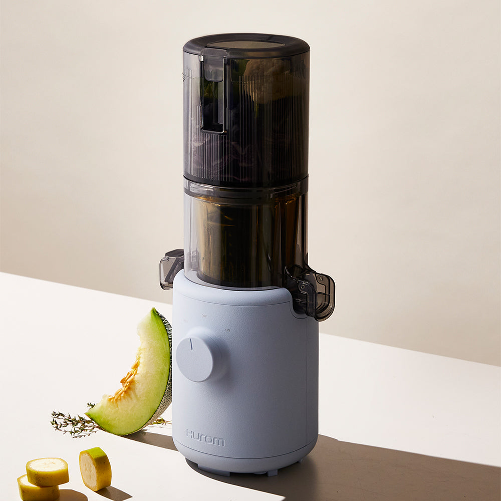 A sky blue Hurom H-310A Slow Juicer on a table, with a melon and some cut bananas beside it.