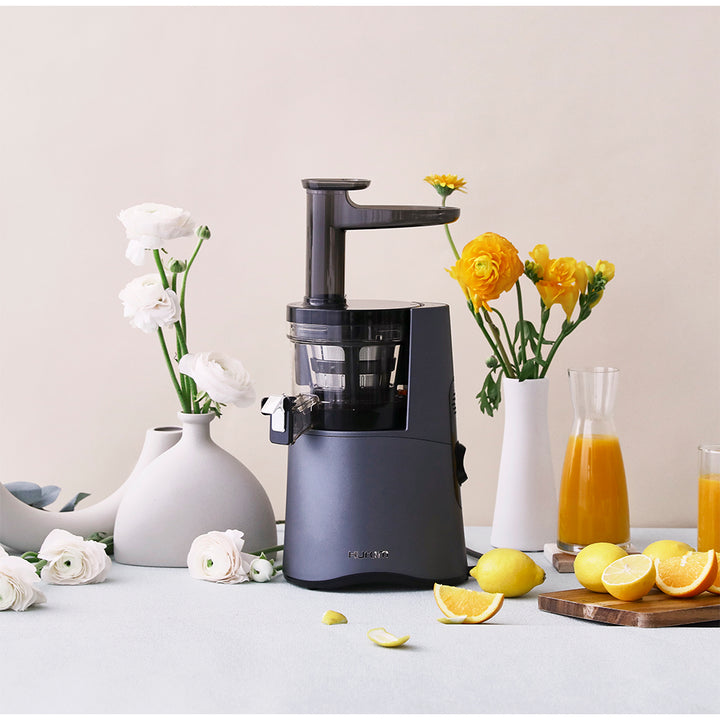 A HA-2600 midnight blue slow juicer surrounded by yellow and white flowers and lemons with a few jugs of orange juice beside, on a light beige backdrop