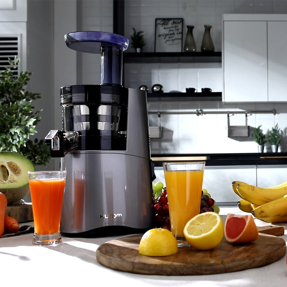 A HA-2600 midnight blue slow juicer with grapes, bananas, lemons and grapefruits surrounding it, with a cup of grapefruit and a cup of lemon juice