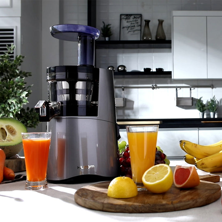 A HA-2600 midnight blue slow juicer with grapes, bananas, lemons and grapefruits surrounding it, with a cup of grapefruit and a cup of lemon juice