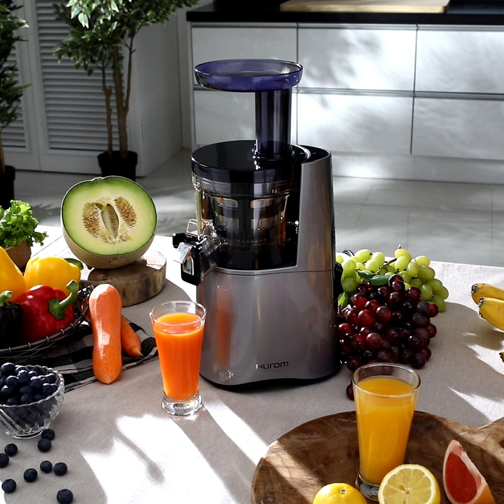 A Hurom HA-2600 midnight blue slow juicer with lots of fruits and vegetables surrounding it including a melon, capsicums, carrots, grapes, lemons and more, with 2 cups of freshly squeezed juices